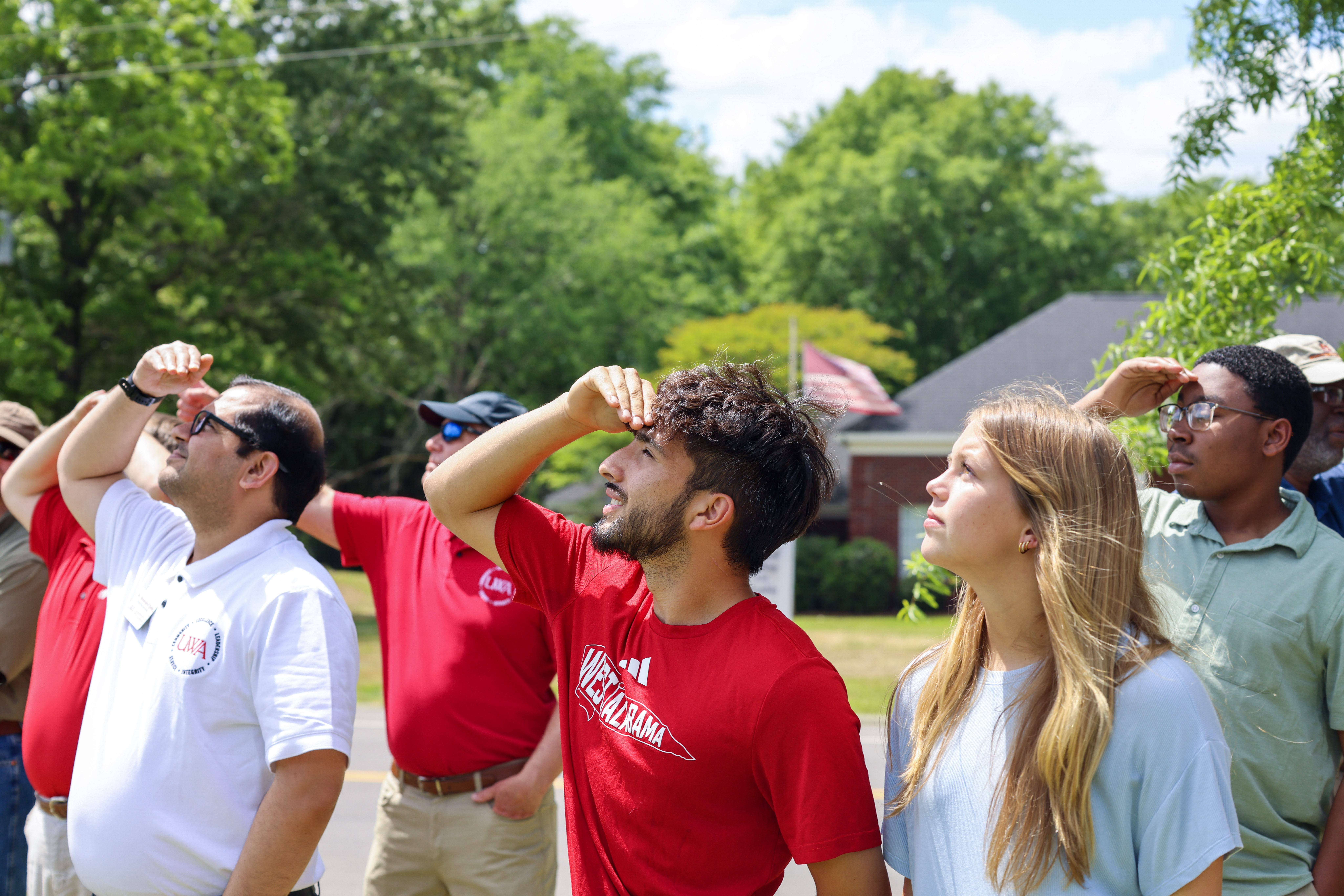 Group of students and instructors looking up outdoors, shielding eyes with hand.